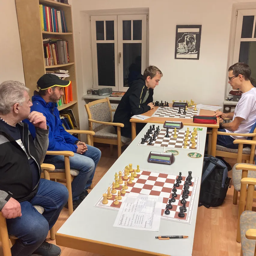 Vier Männer spielen Schach in einem Raum mit Holzböden und Bücherregalen.