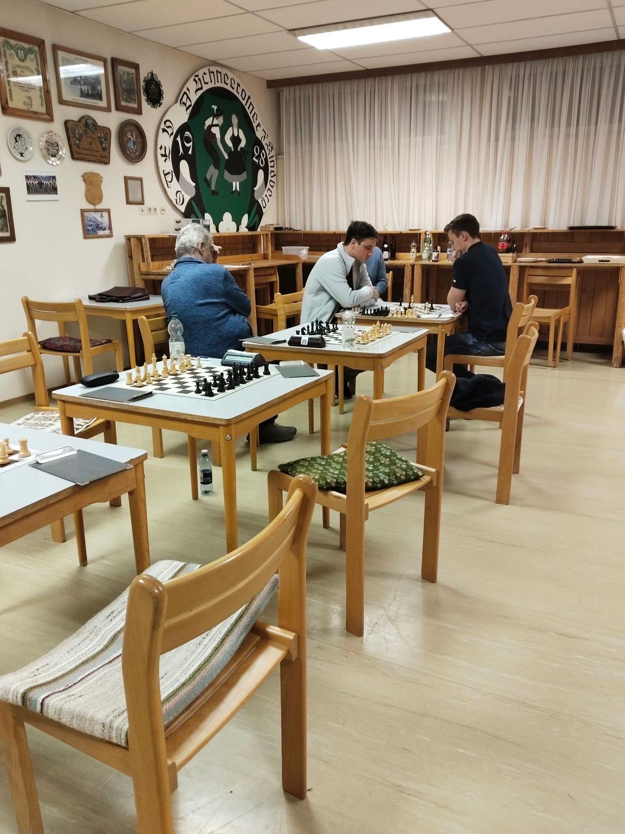 Vier Männer spielen Schach in einem Raum mit Holzstühlen und Tischen.