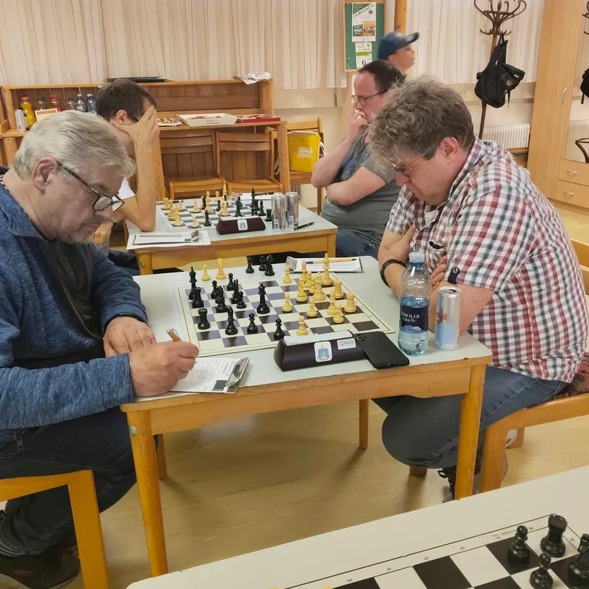 Fünf Männer spielen Schach in einem Raum mit Holzböden und weißen Wänden.