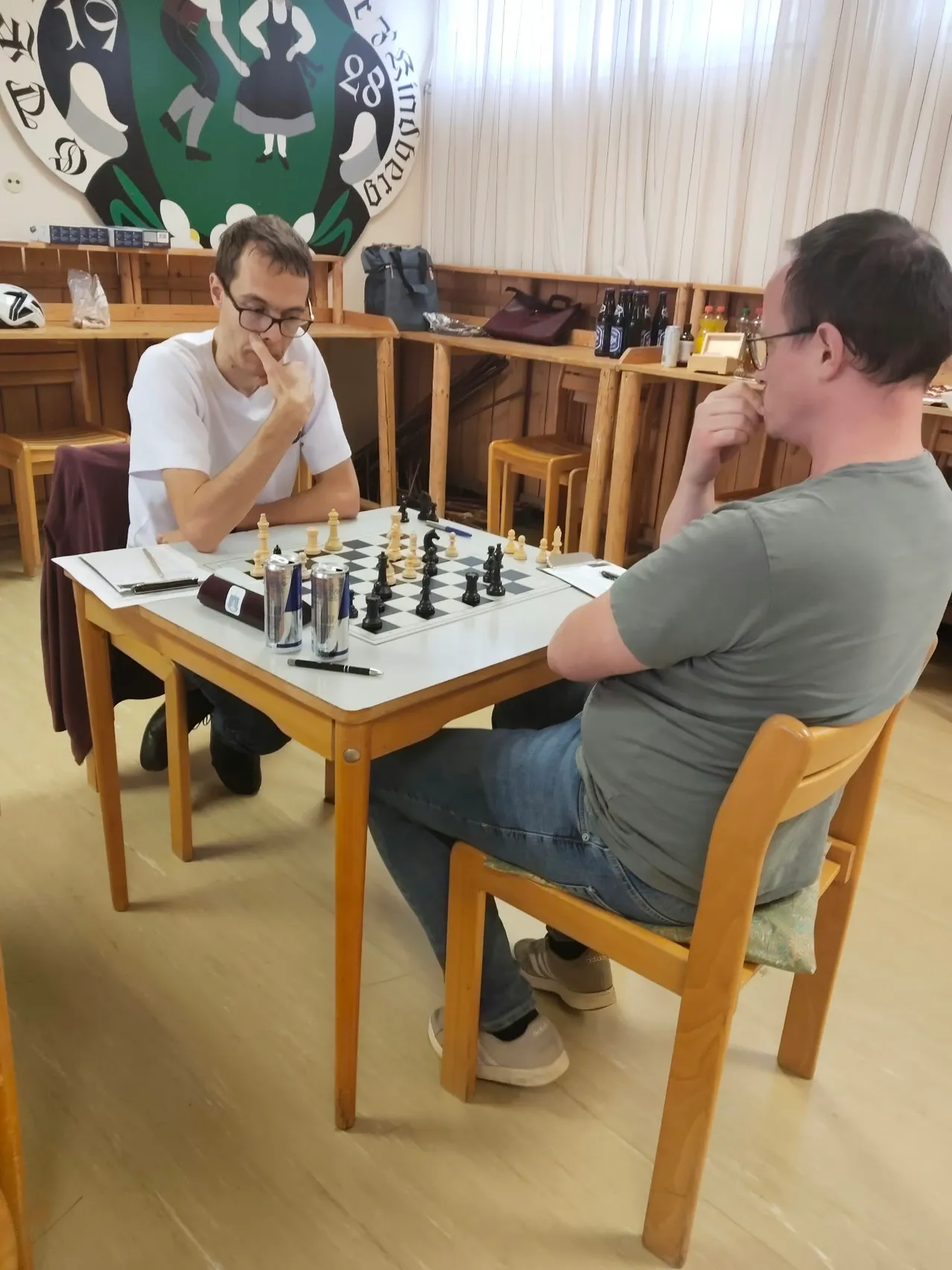 Zwei Männer spielen Schach in einem Raum. Der Mann auf der linken Seite trägt eine Brille und hat seine Hand am Kinn. Der Mann auf der rechten Seite trägt eine Uhr und hat seine Hand am Kinn. Auf dem Tisch vor ihnen steht ein Schachbrett.