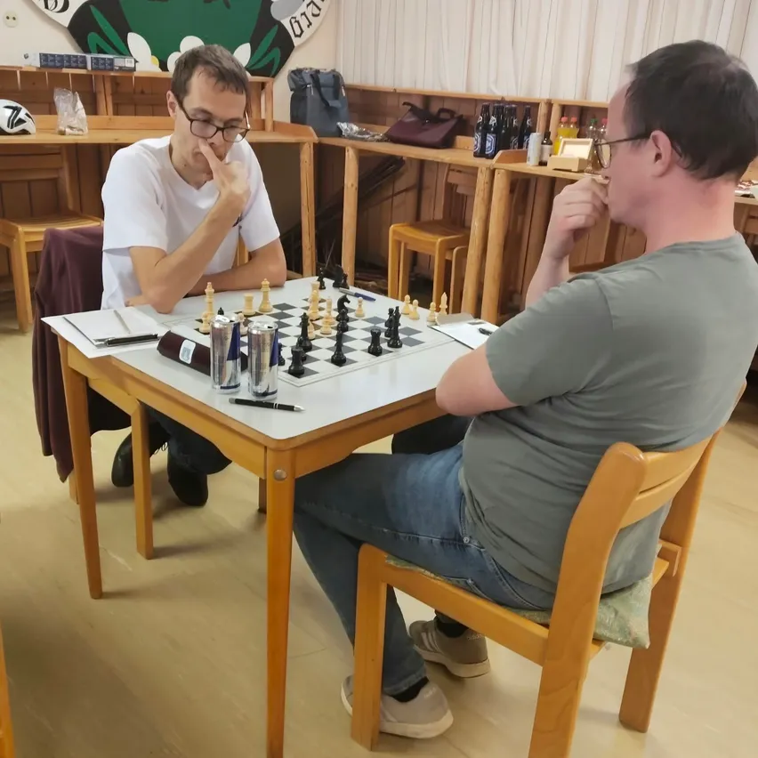 Zwei Männer spielen Schach in einem Raum. Der Mann auf der linken Seite trägt eine Brille und hat seine Hand am Kinn. Der Mann auf der rechten Seite trägt eine Uhr und hat seine Hand am Kinn. Auf dem Tisch vor ihnen steht ein Schachbrett.