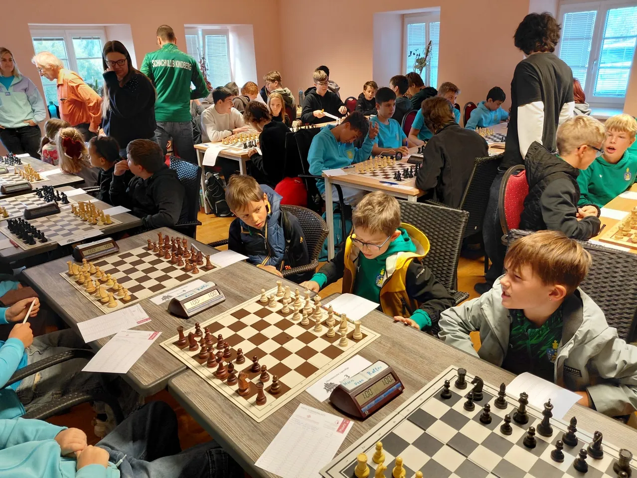 Eine Gruppe von Kindern und Erwachsenen spielt Schach in einem Raum. Die Kinder sitzen an Tischen mit Schachbrettern und -figuren, während Erwachsene um sie herum stehen.