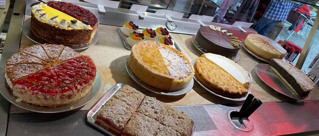 Eine Vitrine mit verschiedenen Kuchen und Gebäck, darunter ein Zitronen-Käsekuchen, ein roter Beerenkuchen und ein Schokoladenkuchen, mit einem Tresen davor.