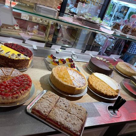Eine Bäckerei-Auslage mit verschiedenen Kuchen auf Tellern. Ein gelber Kuchen mit roten Beeren, ein Schokoladenkuchen und ein Käsekuchen. Davor steht ein Tablett mit mehreren Kuchenstücken darauf.