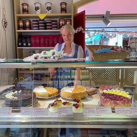 Eine Frau steht hinter einer Glasvitrine in einer Bäckerei voller verschiedener Kuchen. Hinter ihr befinden sich Regale mit Kaffeedosen und Produkten. Draußen vor der Bäckerei sind rosa Blumen und Körbe zu sehen.