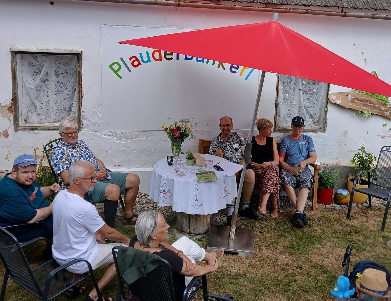 Eine Gruppe von Menschen sitzt um einen Tisch in einem Garten, wobei ein roter Regenschirm Schatten spendet. Die Wand hinter ihnen hat farbenfrohe Schrift.