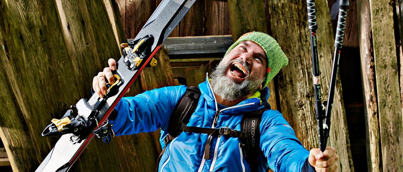 Ein bärtiger Mann mit grüner Mütze hält Skier und trägt einen blauen Mantel. Er lächelt und steht vor einer Holzwand.