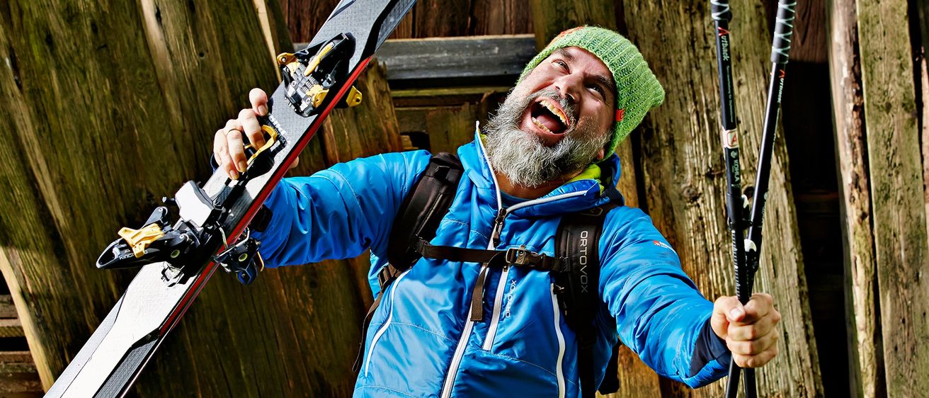 Ein bärtiger Mann mit grüner Mütze hält Skier und trägt einen blauen Mantel. Er lächelt und steht vor einer Holzwand.