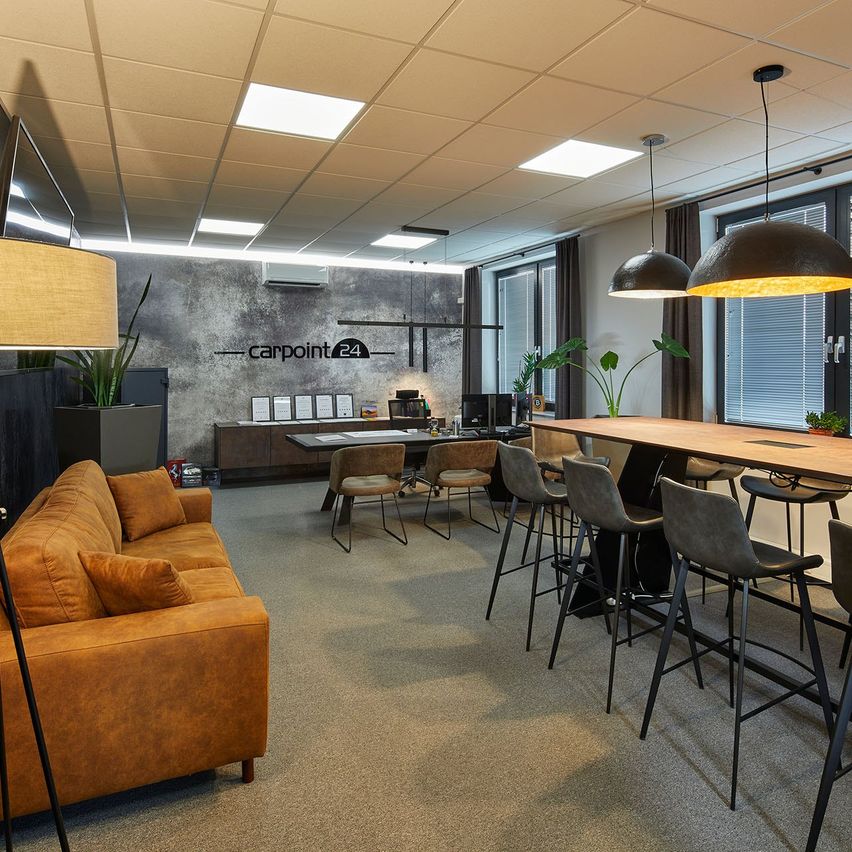 Ein Büroinnenraum mit einer Couch, Stühlen, einem langen Tisch und Pflanzen. Ein Schild an der Wand steht 'carpoint 24'. Es gibt große Fenster mit Jalousien, Lampen und eine Decke mit Lichtern.