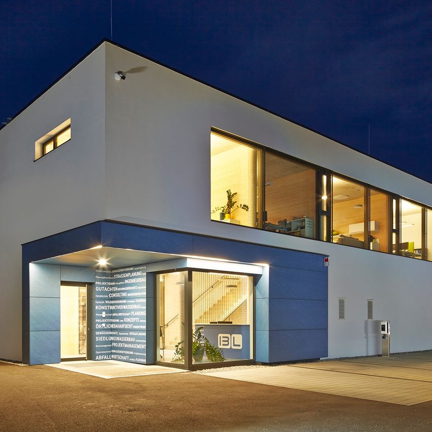 Ein modernes Gebäude mit blauen Akzenten und Glasfenstern ist bei Nacht beleuchtet. Der Eingang hat ein beleuchtetes Schild und eine Pflanze in einem Topf.