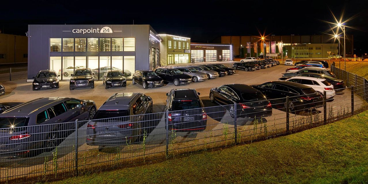 Ein Autohaus bei Nacht mit mehreren Autos draußen geparkt. Das Gebäude hat große Glasfenster und ein Schild, das 'int 24' steht. Die Autos sind in Reihen geparkt, mit einem Metallzaun, der sie von der Straße trennt.