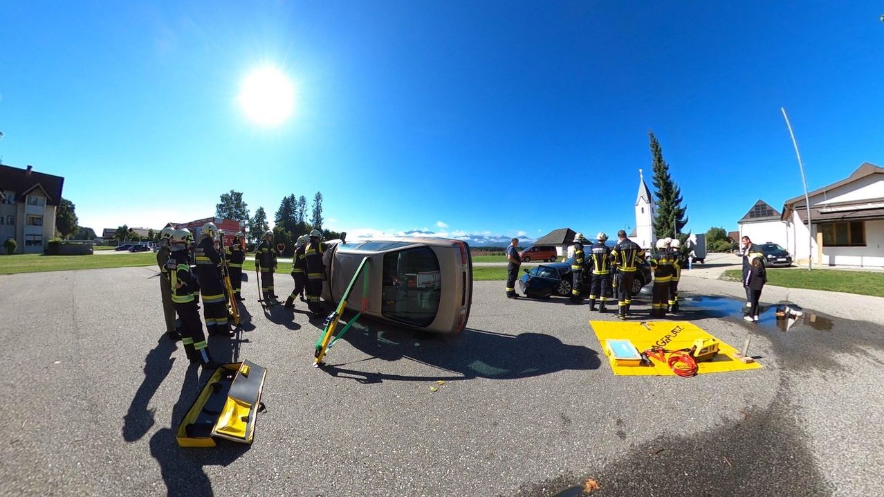 Feuerwehrleute stehen um ein umgekipptes Auto an einem sonnigen Tag, mit Werkzeugen und Ausrüstung in der Nähe.