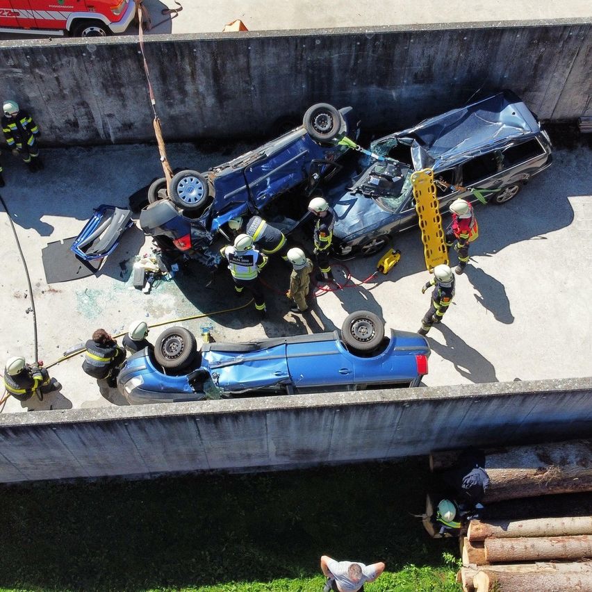 Rettungskräfte kümmern sich um ein umgekipptes blaues Auto auf einer Betonfläche. In der Nähe ist ein weiteres blaues Auto umgekippt, dessen Front stark beschädigt ist. Ein Feuerwehrwagen ist im Hintergrund geparkt.
