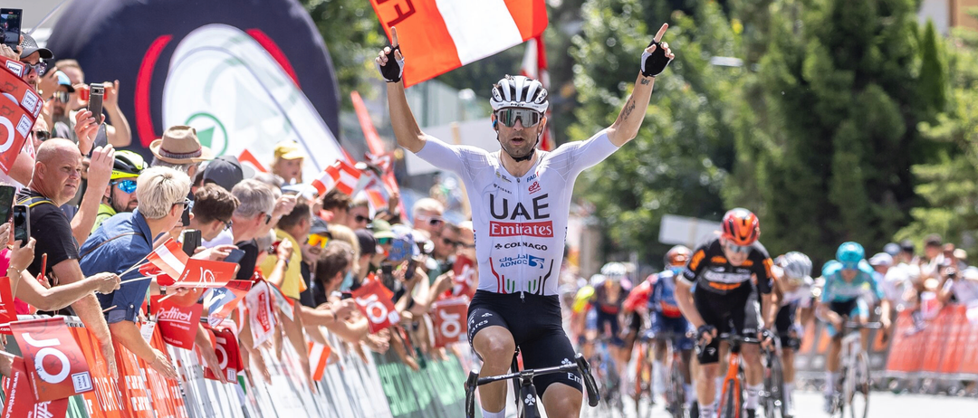 Ein Radfahrer in einem UAE Emirates-Teamtrikot fährt siegreich mit erhobenen Armen, angefeuert von Zuschauern. Hinter ihm folgt eine Gruppe von Radfahrern.