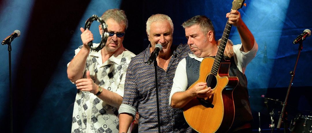 Drei erwachsene Männer treten auf der Bühne auf. Einer singt in ein Mikrofon, während ein anderer die Gitarre spielt und der dritte eine Rassel spielt.