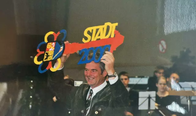 Ein Mann in einem Anzug hält einen großen Schlüssel mit den Worten Stadt 2000 über seinem Kopf. Hinter ihm scheint eine Gruppe von Menschen Instrumente zu spielen.