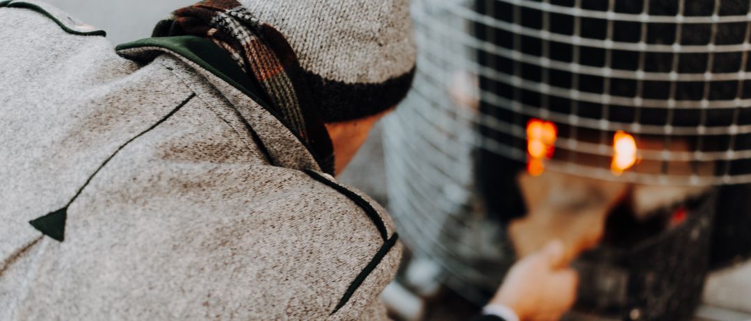 Eine Person in einem grauen Mantel und Hut fügt einem Feuer in einem Metallofen Holz hinzu.