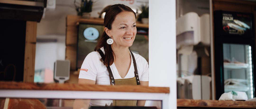 Eine Frau in einem weißen T-Shirt und einer braunen Schürze steht hinter einem Tresen, lächelt und schaut in die Kamera. Sie hat einen weißen Ohrring und befindet sich in einem Raum mit einem Holztresen und Regalen.