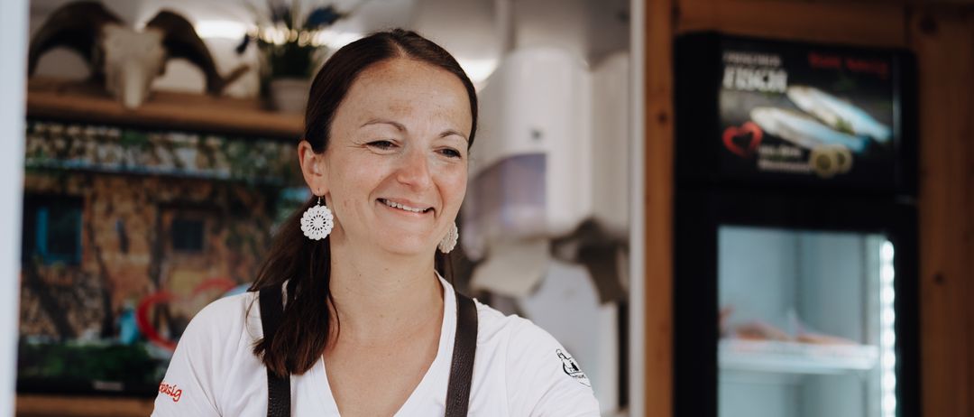 Eine Frau in einem weißen T-Shirt und einer braunen Schürze steht hinter einem Tresen, lächelt und schaut etwas an. Dahinter ist ein Kühlschrank zu sehen, und ein Regal hält verschiedene Gegenstände.