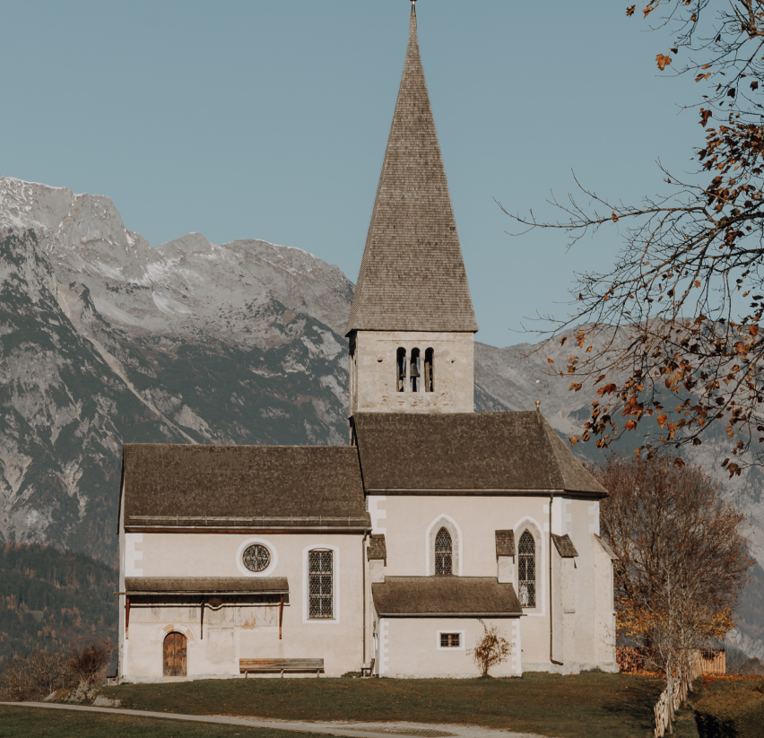 Eine kleine Kirche mit einem Kirchturm und einer Wetterfahne steht in einem grünen Tal mit einer Bank davor und verschneiten Bergen im Hintergrund.