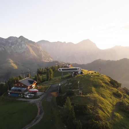 Eine Skiliftstation befindet sich auf einem grünen Hügel unter einem goldenen Sonnenuntergang mit Bergen im Hintergrund.