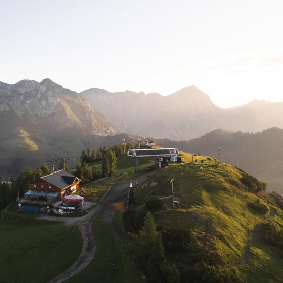 Eine Skiliftstation befindet sich auf einem grünen Hügel unter einem goldenen Sonnenuntergang mit Bergen im Hintergrund.