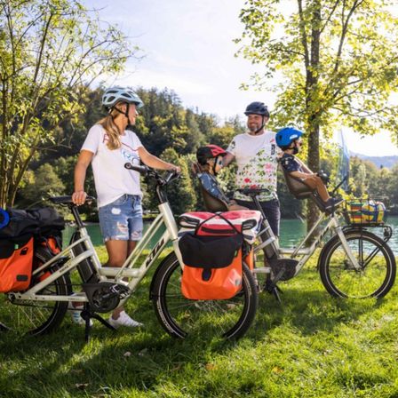 Eine Familie steht mit ihren Fahrrädern in der Nähe eines Sees. Die Frau vorne trägt einen Helm. Der Mann hinter ihr hat ein Blumenhemd und ein Kind auf seinem Fahrrad. Ein weiteres Kind sitzt auf dem Hinterrad des Fahrrads des Mannes.