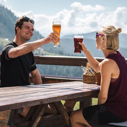 Ein Paar genießt Getränke an einem Holztisch auf einer Bergterrasse. Der Mann hält ein großes Glas Bier, während die Frau ein Glas Limonade in der Hand hat. Hinter ihnen ist ein Blick auf die Berge.