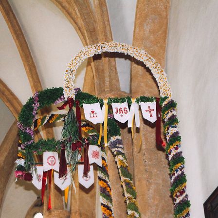 Ein dekorativer Bogen aus Blumen, Bändern und Fahnen, möglicherweise für eine Kirchenzeremonie, hängt an einer Steinmauer.