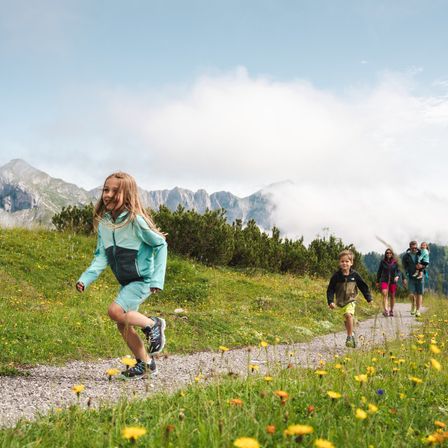 Eine vierköpfige Familie wandert auf einem grasbewachsenen Pfad in den Bergen. Das Mädchen vorne läuft und lächelt. Die Erwachsenen gehen hinter ihr, einer trägt ein Baby.