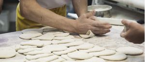 A person in a white shirt and yellow apron is shaping dough on a wooden table with flour sprinkled around. Another hand is visible on the right.