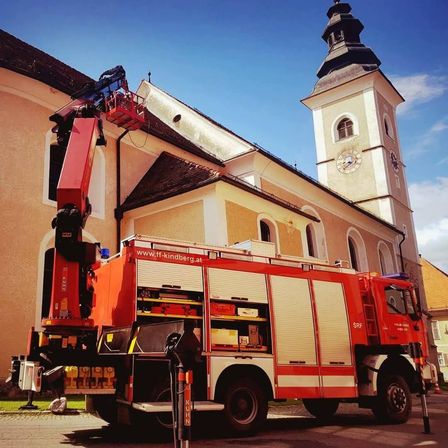 Ein roter Feuerwehrwagen steht vor einer Kirche mit Turm, Uhr und Dachreiter. Ein Kran befindet sich auf dem LKW.