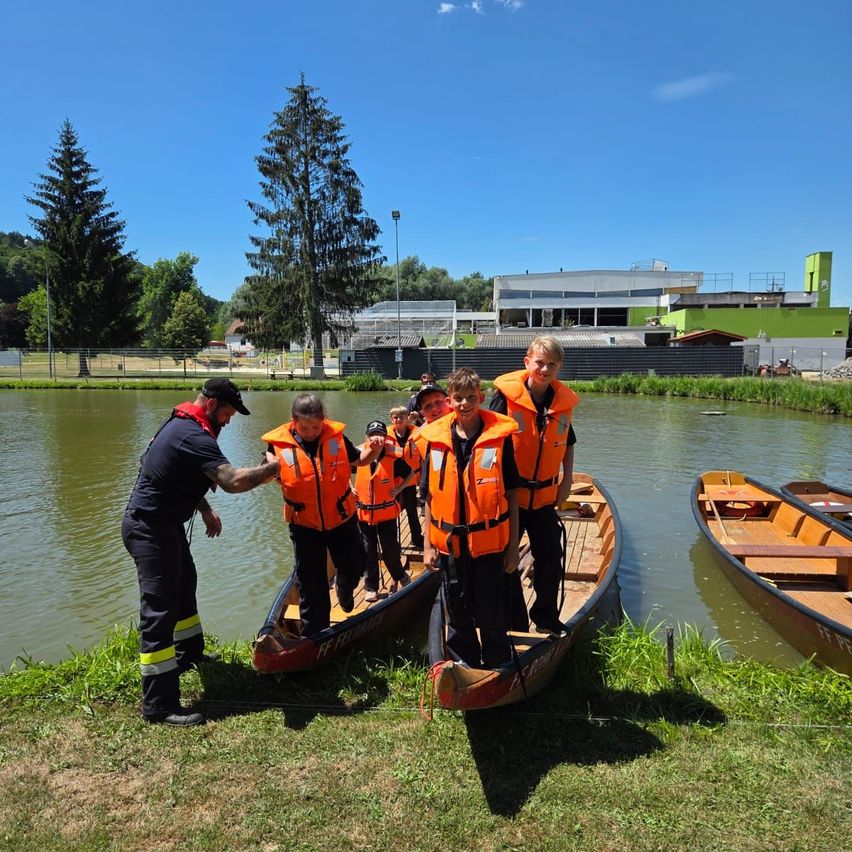 Eine Gruppe von Menschen in orangefarbenen Schwimmwesten steht in Booten am Wasserrand. Ein Mann hilft ihnen.