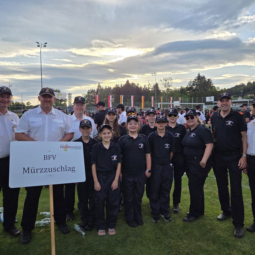 Eine Gruppe von Menschen in schwarzen Uniformen mit weißen Mützen steht auf einem Feld, mit einem Schild, auf dem BFV Murzzuschlag steht.