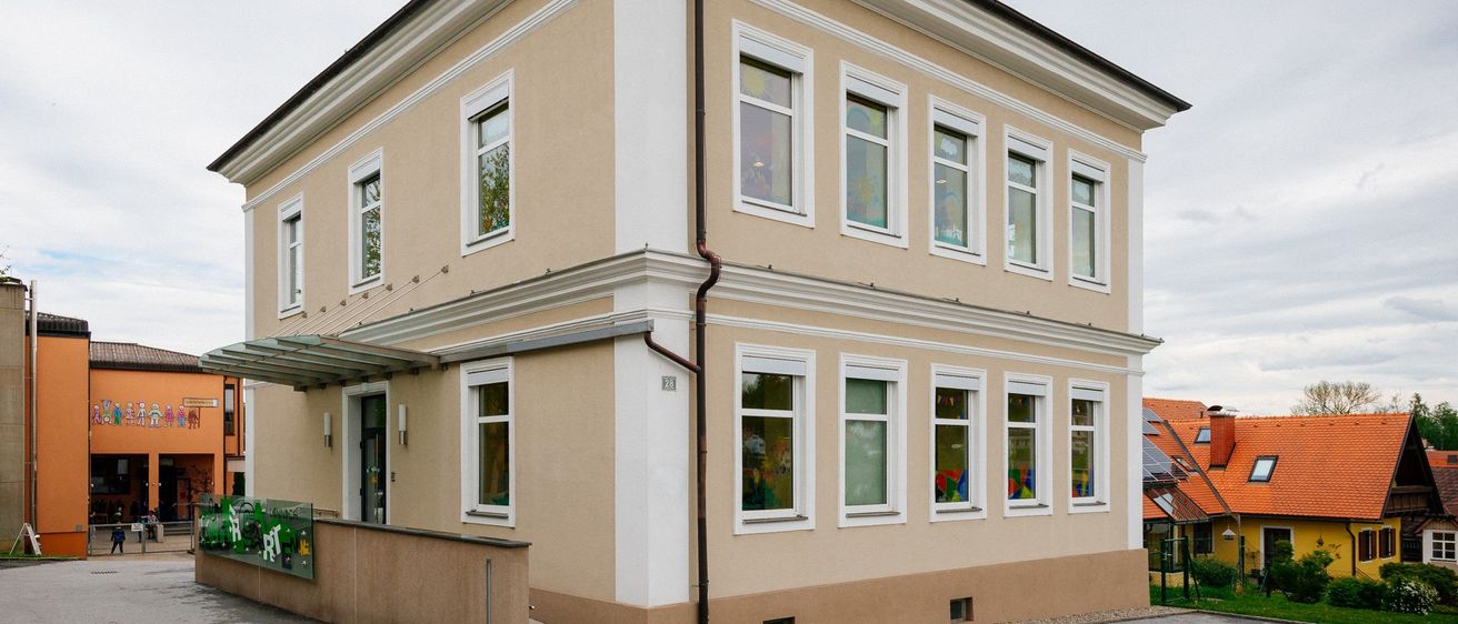 Ein beiger Bau mit weißer Einfassung und vielen Fenstern steht in einer Stadt, mit einem kleinen Vordach und einem Schild. Ein orangefarbenes Haus mit Solarpanelen befindet sich in der Nähe.