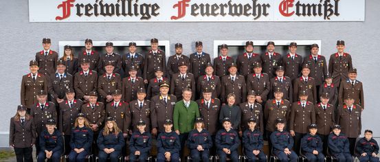 Gruppenfoto der Freiwilligen Feuerwehr Ettlingen mit uniformierten Mitgliedern und Offizieren, die vor einem Schild stehen und sitzen.