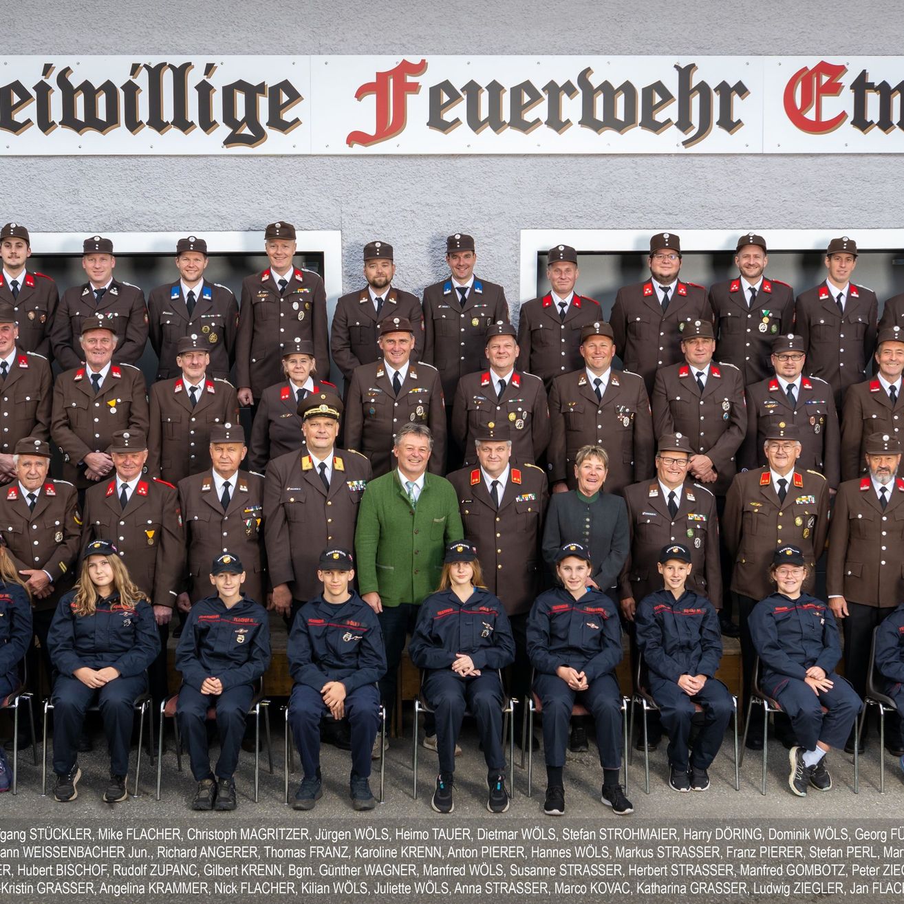 Ein Gruppenfoto der Freiwilligen Feuerwehr Ettlingen, mit Feuerwehrleuten in Uniform, einigen Personen in Zivilkleidung, stehend und sitzend vor einem Gebäude mit einem Schild, das 'Freiwillige Feuerwehr Ettlingen' besagt.