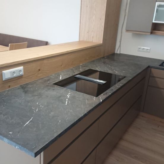 Eine moderne Küche mit einer grauen Marmorarbeitsplatte und einem quadratischen Induktionskochfeld. Die Arbeitsplatte erstreckt sich über Holzschränke, und die Wand ist weiß gestrichen.
