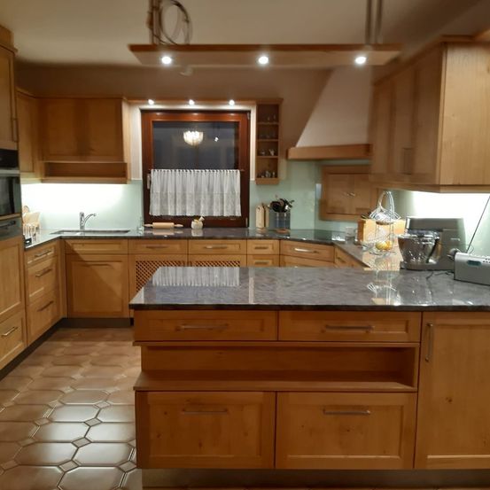 Eine Küche mit Holzschränken, Granitarbeitsplatte und moderner Beleuchtung. Sie verfügt über ein Waschbecken, ein Fenster mit weißen Vorhängen und Geräte.