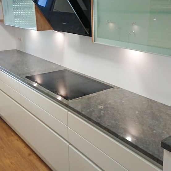 Moderne Küche mit Granitarbeitsplatte, weißen Schränken und einem eleganten Induktionskochfeld unter einer Glasvitrine. Die Wände sind weiß und der Boden besteht aus Holz.