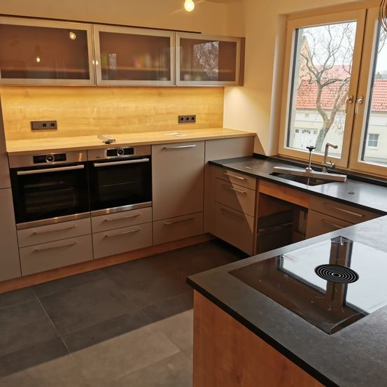 Moderne Küche mit Holzschränken, Glastüren und Granitarbeitsplatte. Zwei Öfen und ein Waschbecken mit Wasserhahn. Ein großes Fenster bietet einen Blick auf einen Baum.