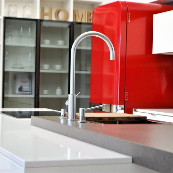 Eine moderne Küche mit einem roten Kühlschrank und einem silbernen Wasserhahn. Gläserne Schränke und Regale mit Tassen und Tellern. Ein Schneidebrett liegt auf dem Spülbecken.