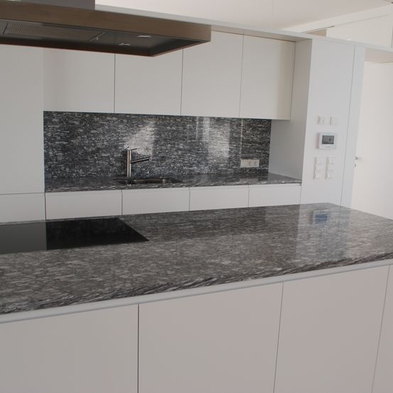 Eine moderne Küche mit weißen Schränken und einer grauen Granitarbeitsplatte. Die Rückwand besteht aus grauen Fliesen, und es gibt ein Edelstahlspülbecken mit einem Wasserhahn. Über der Arbeitsplatte befindet sich eine Dunstabzugshaube.