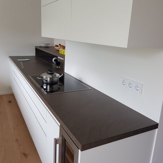 Eine moderne Küche mit weißen Schränken und einer schwarzen Arbeitsplatte. Es gibt einen Gasherd mit einem Topf darauf und eine Steckdose an der Wand.