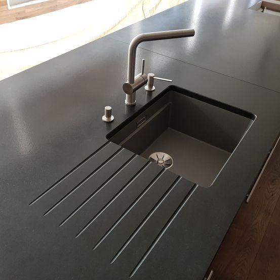Eine moderne schwarze Küchenarbeitsplatte mit Spüle, Edelstahlhahn und Abtropffläche. Die Spüle hat ein modernes Design mit Rillen auf der Arbeitsplatte.