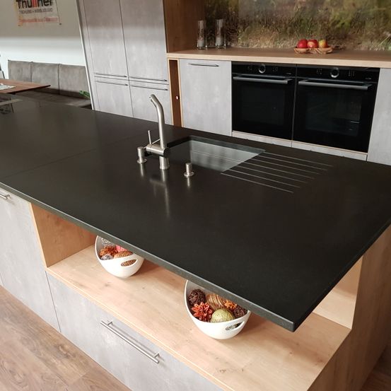 Eine moderne Küche mit einer großen schwarzen Granitarbeitsplatte, einem Waschbecken mit Wasserhahn und zwei Schüsseln mit verschiedenen Früchten und Gemüse. Hinter der Arbeitsplatte befindet sich ein Schrank mit Herd und Backofen sowie ein Tisch mit einer Schüssel mit Obst.
