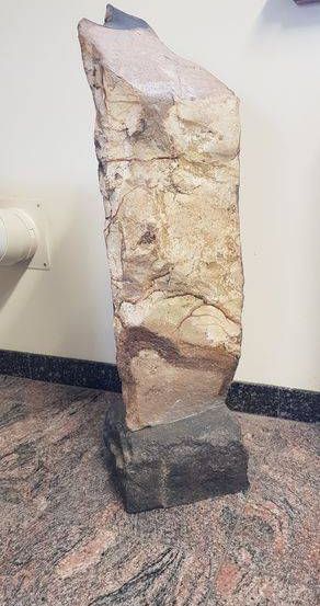 Eine große, raue Steinskulptur steht auf einem Marmorsockel. Sie hat eine natürliche, erdige Textur mit braunen und beigen Farbtönen. Die Skulptur steht an einer weißen Wand.