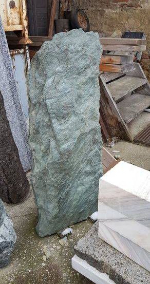 Ein großer grüner Felsblock liegt auf einem Betonboden, neben einem Haufen Holzplanken. Verschiedene Steinplatten und eine Holztreppe sind im Hintergrund.