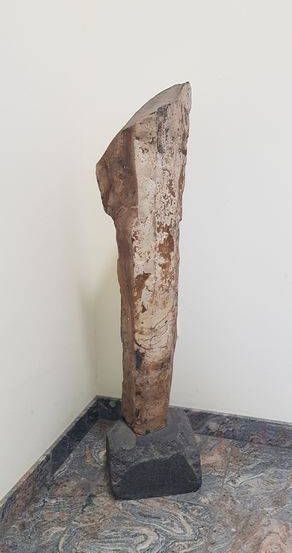 Eine Steinskulptur steht in einer Ecke, gestützt von einem grauen Block auf einem fleckigen Marmorboden gegen eine weiße Wand.