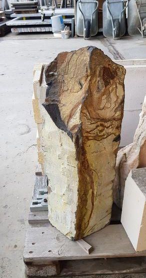 Ein großer Steinblock mit komplizierten Mustern, der auf einem Holzpalette in einer Werkstatt ruht. Im Hintergrund sind Eimer und Schubkarren zu sehen.